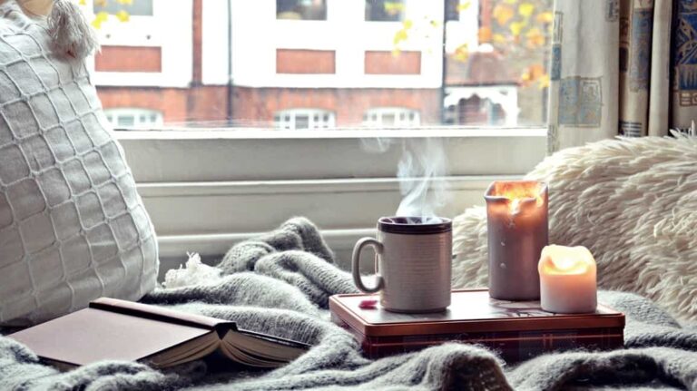Candles and coffee on a bedside table on top of a cozy bed with an open book.